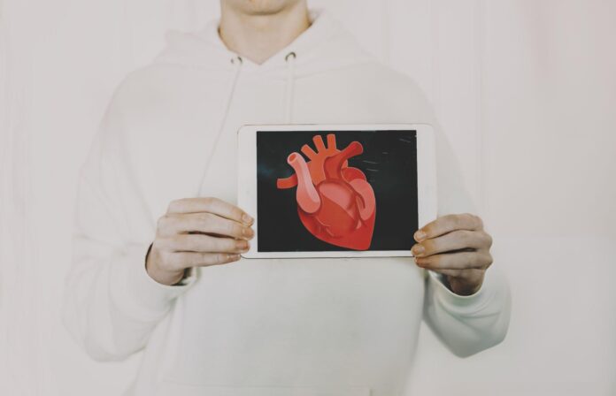 Person me bluzë të bardhë me kapuç që mban një tablet ku shfaqet një ilustrim digjital i zemrës njerëzore. Zemra paraqitet me detaje anatomike në nuanca të kuqe dhe rozë, mbi sfond të zi, duke simbolizuar ndërthurjen mes teknologjisë dhe edukimit për sëmundjet e zemrës si perikarditi.