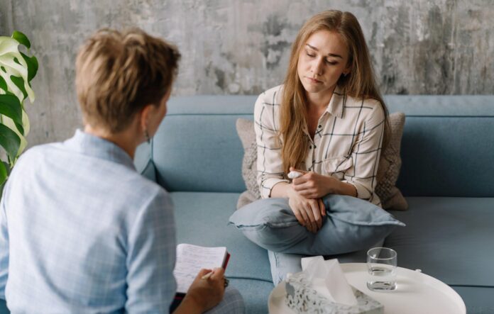 Dy persona në një ambient të qetë terapeutik, ku njëri ndan ndjenja personale ndërsa tjetri merr shënime. Personi në divan blu mban një jastëk dhe letërthithëse, duke reflektuar emocione të thella. Atmosfera sugjeron një seancë psikoterapie, e përshtatshme për trajtimin e çrregullimeve të shëndetit mendor si çrregullimi bipolar. Elementet si kutia e letërthithëseve, gota e ujit dhe bima dekorative përforcojnë ndjesinë e mbështetjes dhe sigurisë.