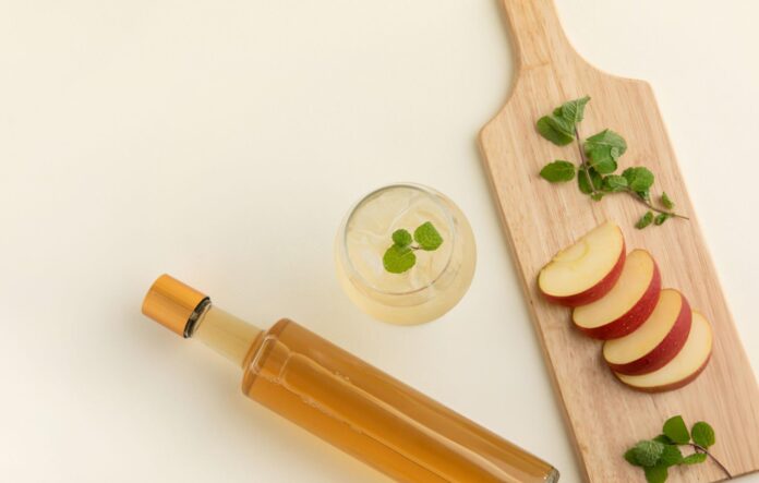 Kompozim i kuruar me një shishe me lëng ngjyrë qelibar, një gotë me pije të freskët me mente dhe akull, dhe një dërrasë druri me feta mollë të kuqe dhe gjethe menteje. Sfondi krem i çelët thekson freskinë dhe natyralitetin e përbërësve, duke sugjeruar përdorimin e mollës dhe uthullës së mollës në receta të shëndetshme ose pije rigjallëruese.